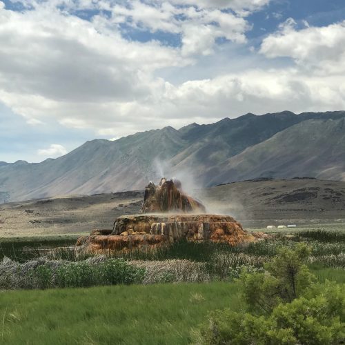 A colorful geothermal feature rises from the ground, surrounded by green grass and mountains under a cloudy sky.