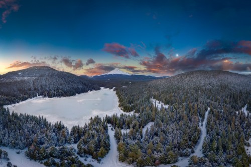 A serene winter landscape featuring a frozen lake surrounded by snow-covered trees and mountains under a colorful sky.