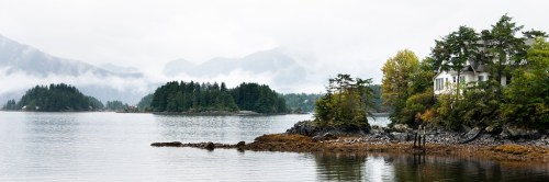 A serene coastal landscape featuring misty mountains, calm waters, and a house surrounded by trees.