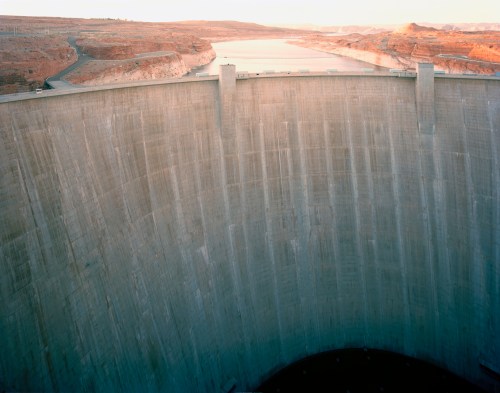Aerial view of a large concrete dam with a river and rocky landscape in the background during sunset.
