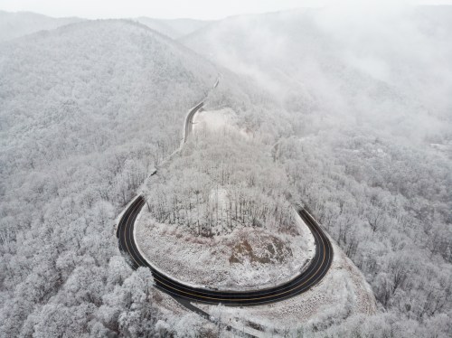 Aerial view of a winding road through snow-covered mountains and trees, shrouded in mist.