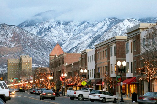 A snowy mountain backdrop frames a charming downtown street lined with shops and festive lights.