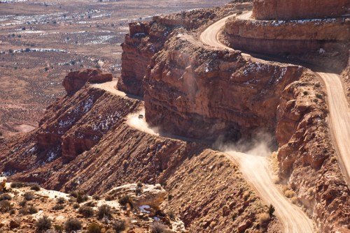 A winding dirt road curves along a rocky cliff in a desert landscape, with dust rising from a vehicle.