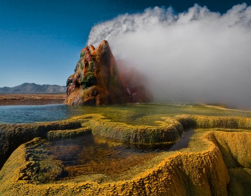 A colorful geothermal spring erupts steam and water, surrounded by vibrant mineral deposits and a clear blue sky.