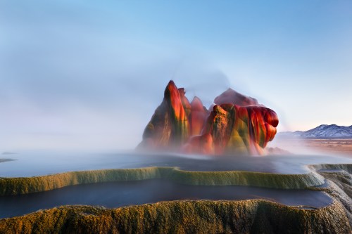 A colorful rock formation emerges from misty water, surrounded by a serene landscape under a clear sky.