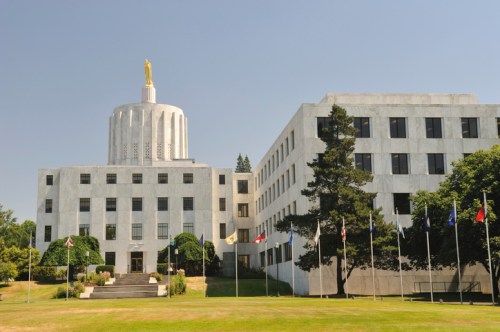Oregon State Capitol building with a gold statue on top, surrounded by trees and flags on a clear day.
