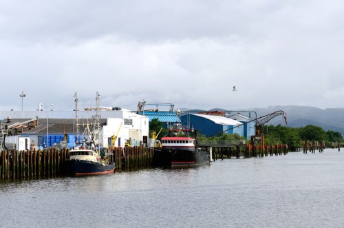 Two fishing boats docked at a harbor with industrial buildings and a cloudy sky in the background.