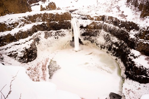 A frozen waterfall cascades into a snow-covered canyon, surrounded by rocky cliffs and a serene, icy landscape.