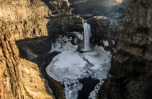 Aerial view of a waterfall cascading into a frozen pool, surrounded by rocky cliffs and a winter landscape.
