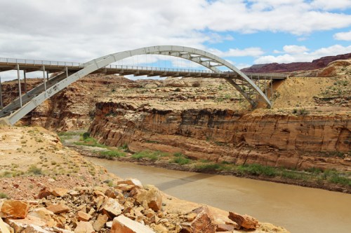 A modern arch bridge spans a river, surrounded by rocky cliffs and a cloudy blue sky.