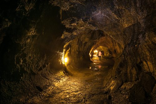 A person walks through a dimly lit cave, with rocky walls and glowing lights illuminating the wet path.
