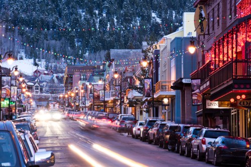 A snowy street lined with festive lights, shops, and parked cars, set against a mountain backdrop.