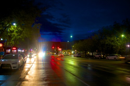 A wet street at night, illuminated by streetlights and car headlights, with trees lining the road.