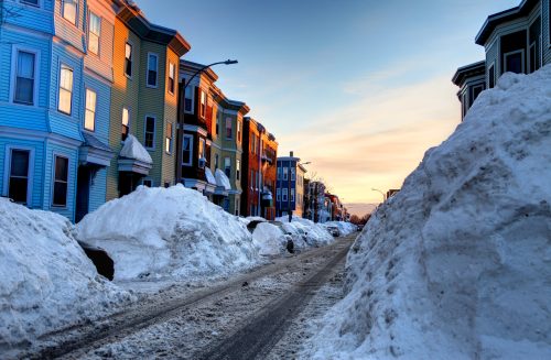Snow-covered street lined with colorful houses at sunset, with large snowbanks on either side.