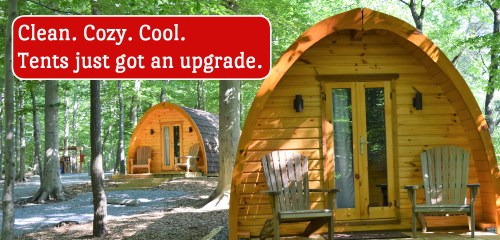 Two wooden glamping cabins nestled in a forest, with a sign reading "Clean. Cozy. Cool. Tents just got an upgrade."