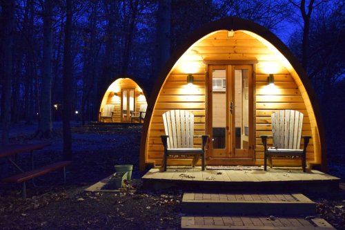 Two cozy wooden cabins with arched roofs, warmly lit at night, surrounded by trees and a picnic table nearby.