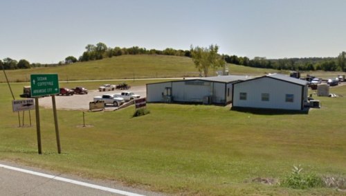 A roadside view of a building with a sign, surrounded by grassy fields and parked vehicles.