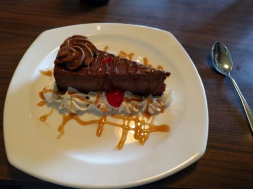 A slice of chocolate cake topped with whipped cream, a cherry, and caramel drizzle on a white plate.
