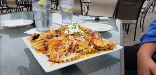 A large plate of loaded waffle fries topped with cheese, sour cream, tomatoes, and green onions, with drinks in the background.