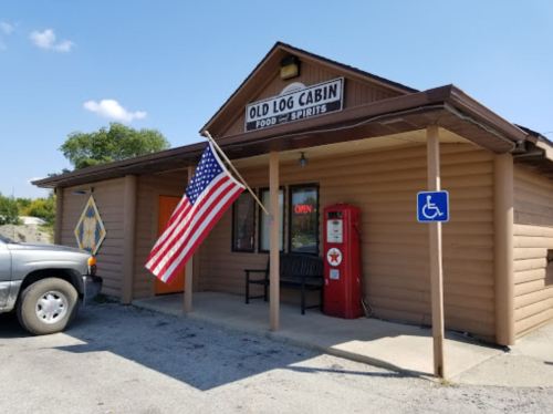 Exterior of Old Log Cabin restaurant with an American flag, a bench, and a vintage soda machine. Clear blue sky above.