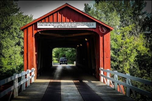 A red covered bridge with a truck entering, surrounded by lush greenery and a clear blue sky.