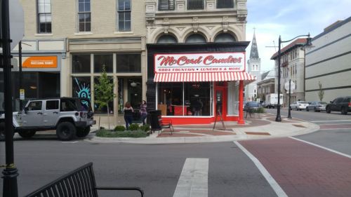 A bright red storefront with a striped awning, featuring "McCosh Candies" and outdoor seating on a city street.