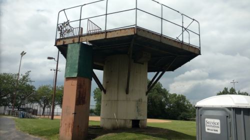 A tall, rusted observation tower on a grassy field, with a portable restroom nearby and cloudy skies above.