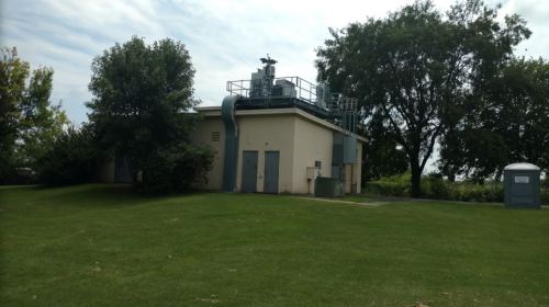 A small utility building with equipment on the roof, surrounded by grass and trees, with a portable restroom nearby.