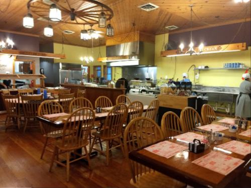 Cozy restaurant interior with wooden tables, chairs, and a visible kitchen area in the background. Warm lighting.