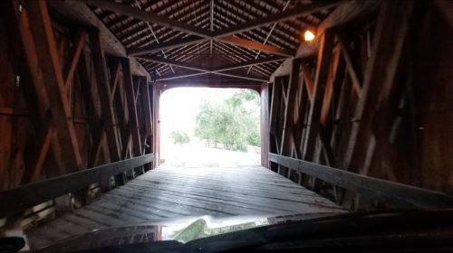 View from inside a covered bridge, showing wooden beams and an opening leading to a grassy area outside.