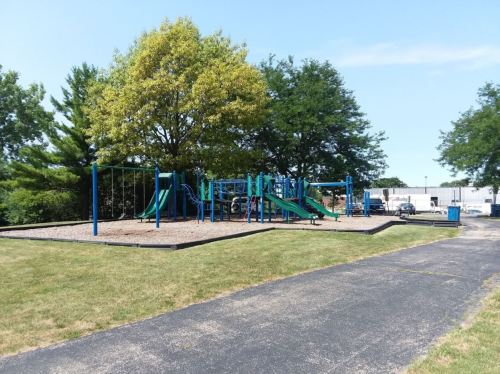 A playground with green slides and swings, surrounded by trees and grass on a sunny day.