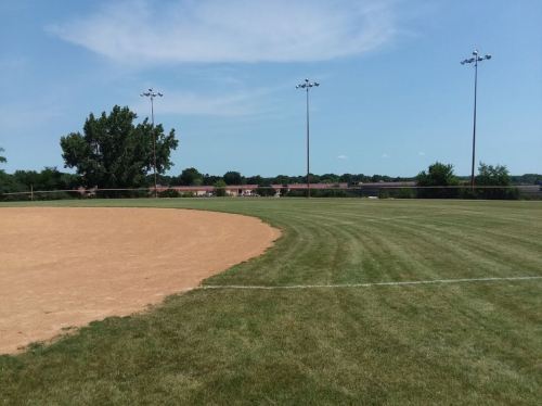 A baseball field with a dirt infield and green grass, under a clear blue sky with a few scattered clouds.