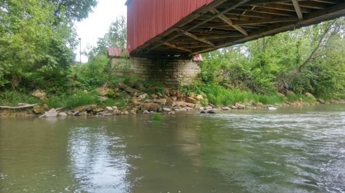 A red covered bridge spans over a calm river, surrounded by lush greenery and rocky riverbanks.