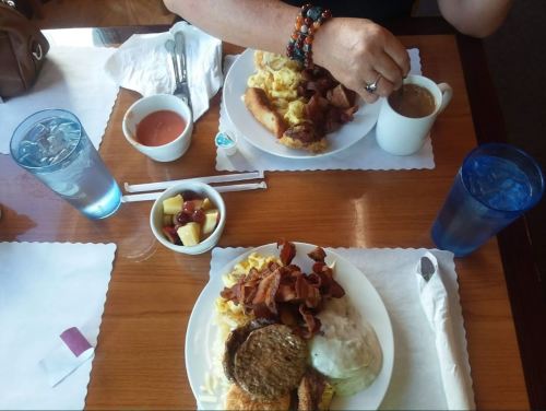 Two plates of breakfast with eggs, bacon, sausage, biscuits, and fruit, alongside cups of coffee and water.