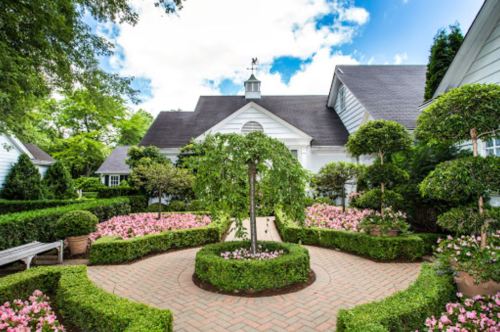 A beautifully landscaped garden with circular pathways, topiary trees, and vibrant pink flowers surrounding a charming house.