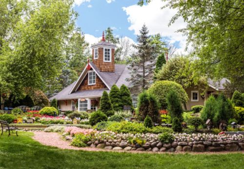 A charming house surrounded by vibrant gardens, colorful flowers, and lush greenery under a blue sky.
