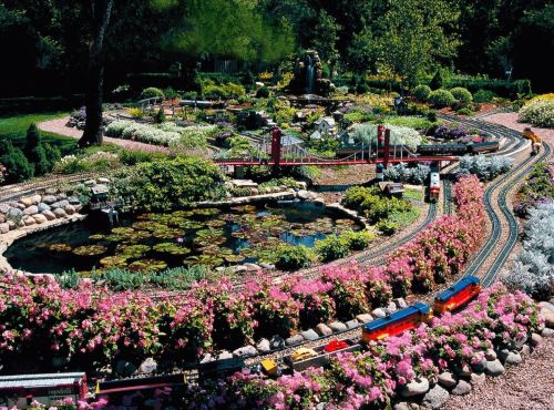A vibrant garden scene featuring a miniature train winding through colorful flowers and a pond.