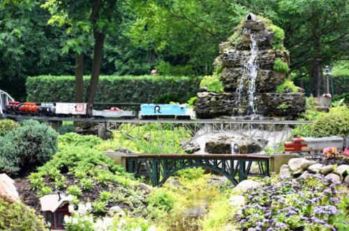 A miniature train travels over a bridge near a waterfall, surrounded by lush greenery and colorful flowers.