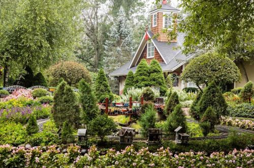 A lush garden with colorful flowers, miniature trains, and a charming house surrounded by trees.