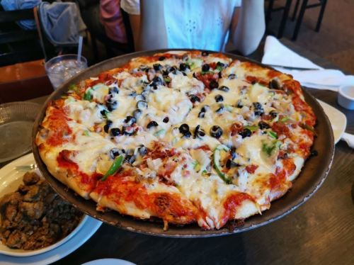 A large pizza topped with cheese, olives, green peppers, and various toppings, served on a round metal tray.