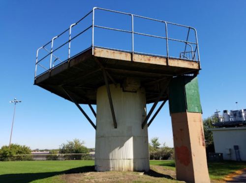 A tall, weathered observation tower with a metal railing, standing on a grassy field under a clear blue sky.
