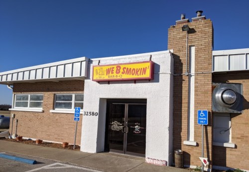 Exterior of a barbecue restaurant with a sign reading "We B Smokin' Bar-B-Q" and a clear blue sky above.
