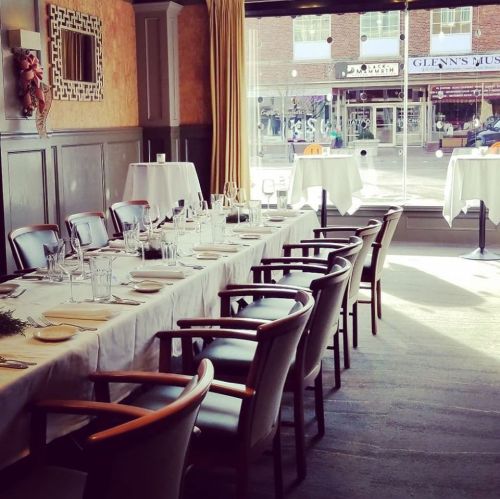 A dining room with a long table set for a meal, featuring elegant chairs and large windows overlooking a street.