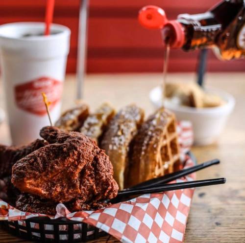 A basket of fried chicken, waffles, and syrup, with a drink in a cup, set on a wooden table.
