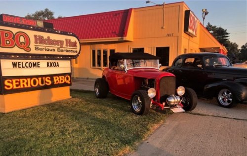 A red vintage car parked in front of a BBQ restaurant with a welcoming sign and another classic car nearby.