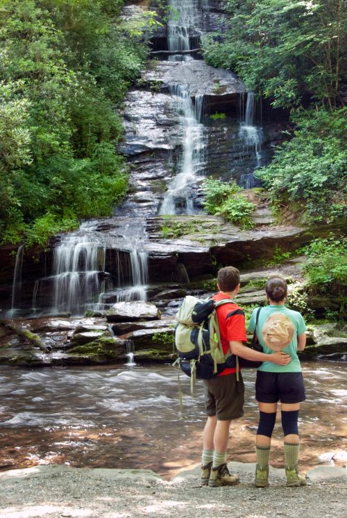 A couple stands by a serene waterfall, surrounded by lush greenery, enjoying nature together.