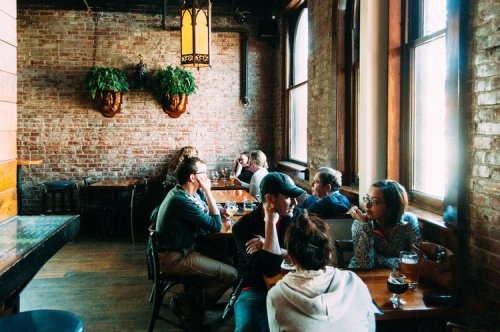 A cozy restaurant interior with people seated at wooden tables, enjoying drinks and conversation by large windows.