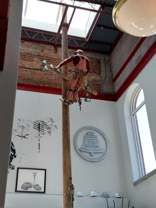 A wooden pole with a figure dressed as a lineman climbing it, inside a building with industrial decor.