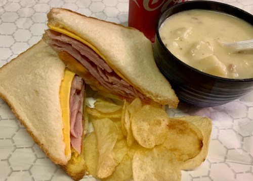 A sandwich with ham and cheese, a bowl of clam chowder, and a side of potato chips, with a soda in the background.