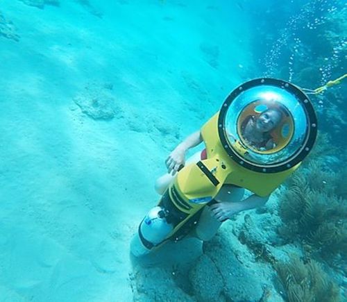 A person in a yellow underwater scooter with a clear dome, exploring the ocean floor surrounded by bubbles and coral.
